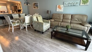 Una sala de estar con sofá, sillón y mesa de centro, un comedor con sillas y una cocina al fondo.