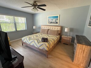 Une chambre à coucher comprenant un lit, deux tables de chevet, un ventilateur de plafond, une fenêtre avec des stores et un tableau encadré accroché au mur.