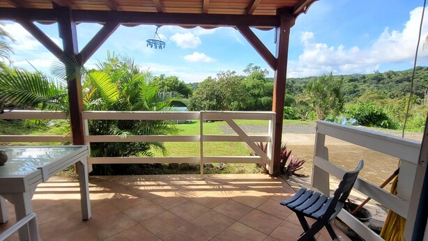 A covered patio with a table and chairs, a view of greenery, and a wooden ceiling structure. - Sofaia Studio: Quiet, relaxing garden view, 2 km from the sulfur baths of Sofaia (Sainte-Rose)