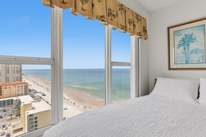 Ein Hotelzimmer mit großem Fenster mit Blick auf einen Strand und den Ozean, einem Bett mit weißer Bettwäsche und einem gerahmten Bild an der Wand.