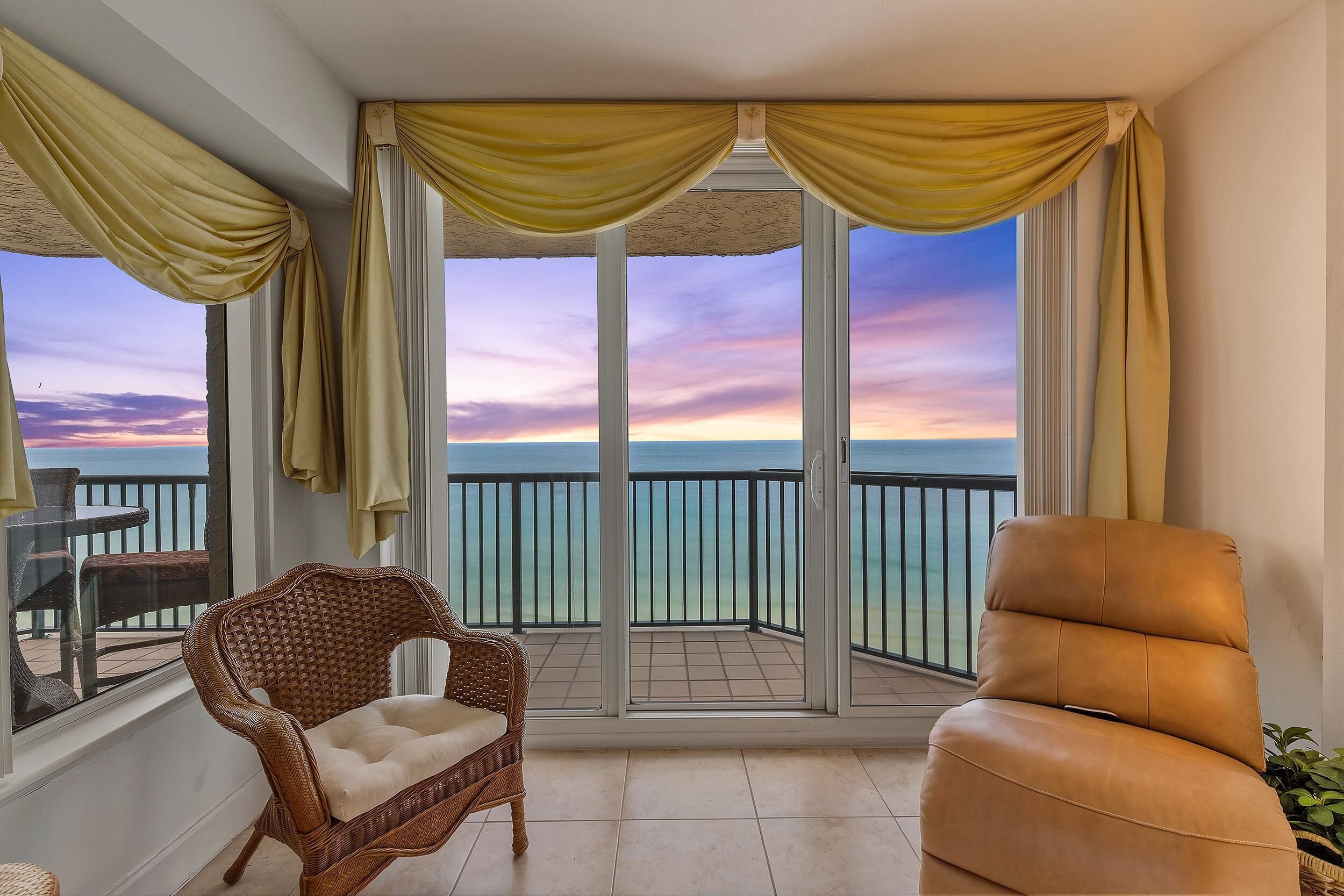 A balcony with a wicker chair, a leather sofa, and a view of the sea at sunset.