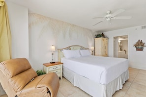 A bedroom with a bed, a chair, a nightstand with a lamp, a ceiling fan, and a wall with a palm tree mural. - Dimucci Towers Ten 1901 (Daytona Beach Shores)