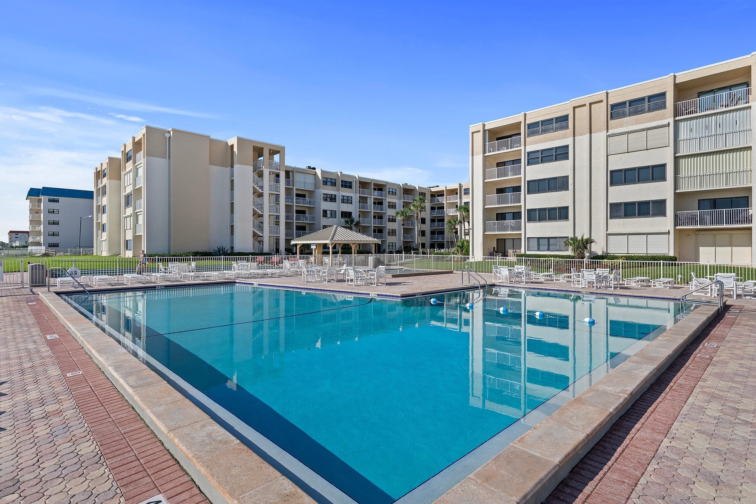 A swimming pool with lounge chairs and a covered seating area, surrounded by a fence.