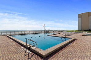 Ein rechteckiger Swimmingpool mit einer weißen Liegefläche, einer Flagge und Blick auf den Strand.