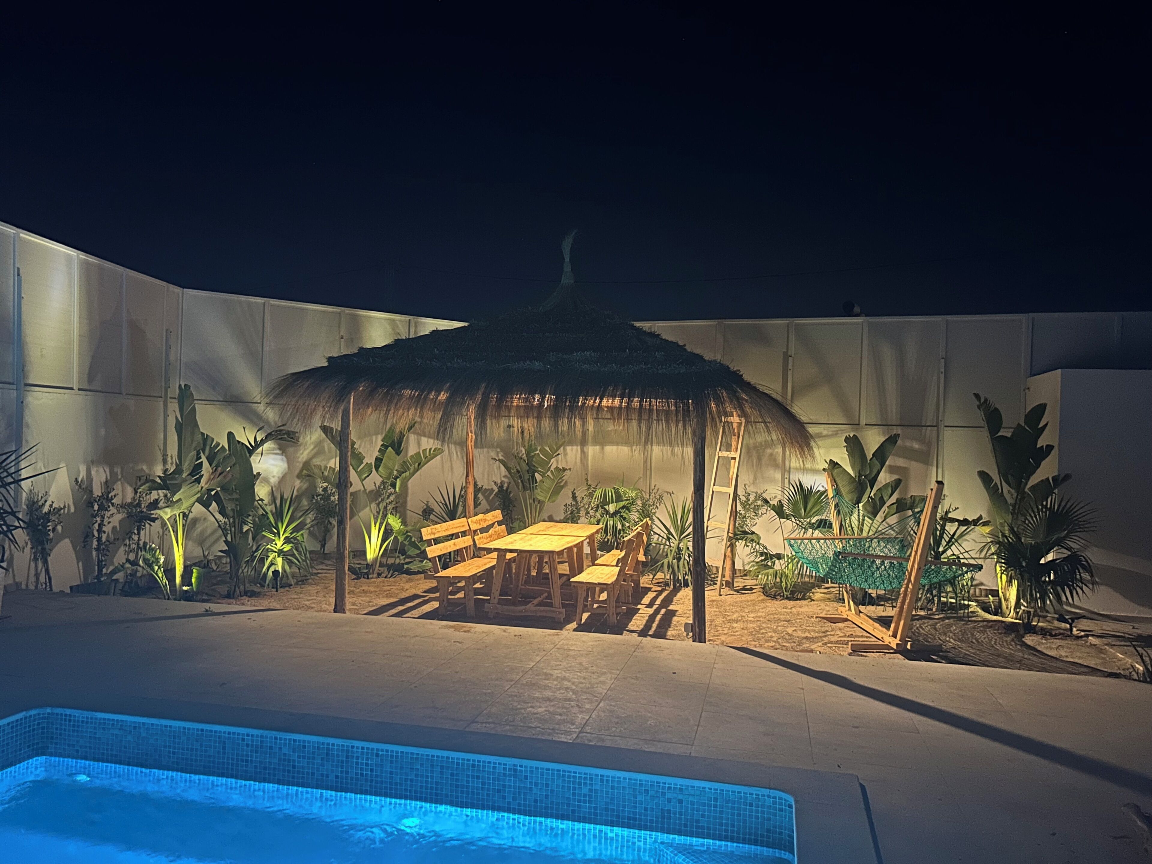 A pool area with a thatched-roof seating area, surrounded by tropical plants.