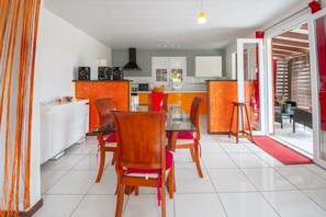 A modern kitchen with white cabinets, a central island, and a dining area with wooden chairs and a glass table. - Maza - Charmante maison pour 4p (Le Lorrain)