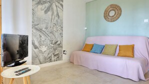 A living room with a television, a round table with remotes, a sofa with cushions, a wall mural, and a wall-mounted decorative item. - Le Cocon de Boucan Canot - À 300 m de la plage (La Possession)