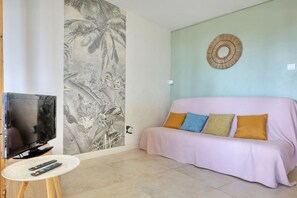 A living room with a television, a round table with remotes, a sofa with cushions, a wall mural, and a wall-mounted decorative item. - Le Cocon de Boucan Canot - À 300 m de la plage (La Possession)