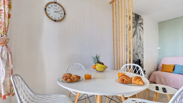 A dining area with a round table, chairs, a clock, and a fruit bowl. - Le Cocon de Boucan Canot - À 300 m de la plage (La Possession)