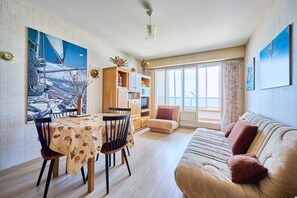 Ein Wohnzimmer mit einer Couch, einem Esstisch mit Stühlen, einem Bücherregal und einem großen Fenster.