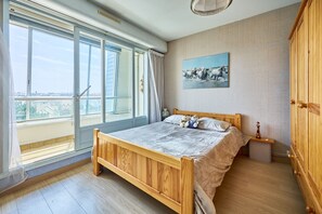 Ein Schlafzimmer mit einem hölzernen Bett, einem Nachttisch, einem Gemälde und Blick durch große Fenster.