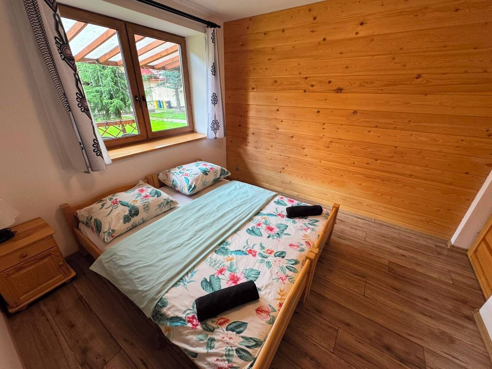 Un dormitorio con piso de madera, una cama con ropa de cama floral, una ventana con cortinas y vistas a la vegetación exterior.