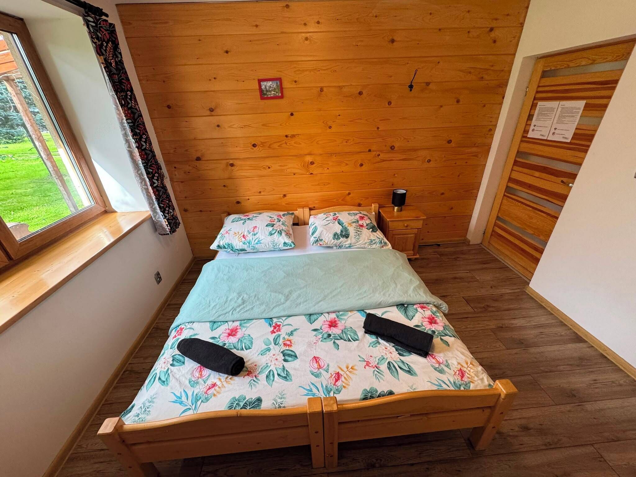 Un lit en bois avec une literie à motifs floraux, dans une pièce dont le plafond est également en bois et qui possède une fenêtre avec des rideaux.