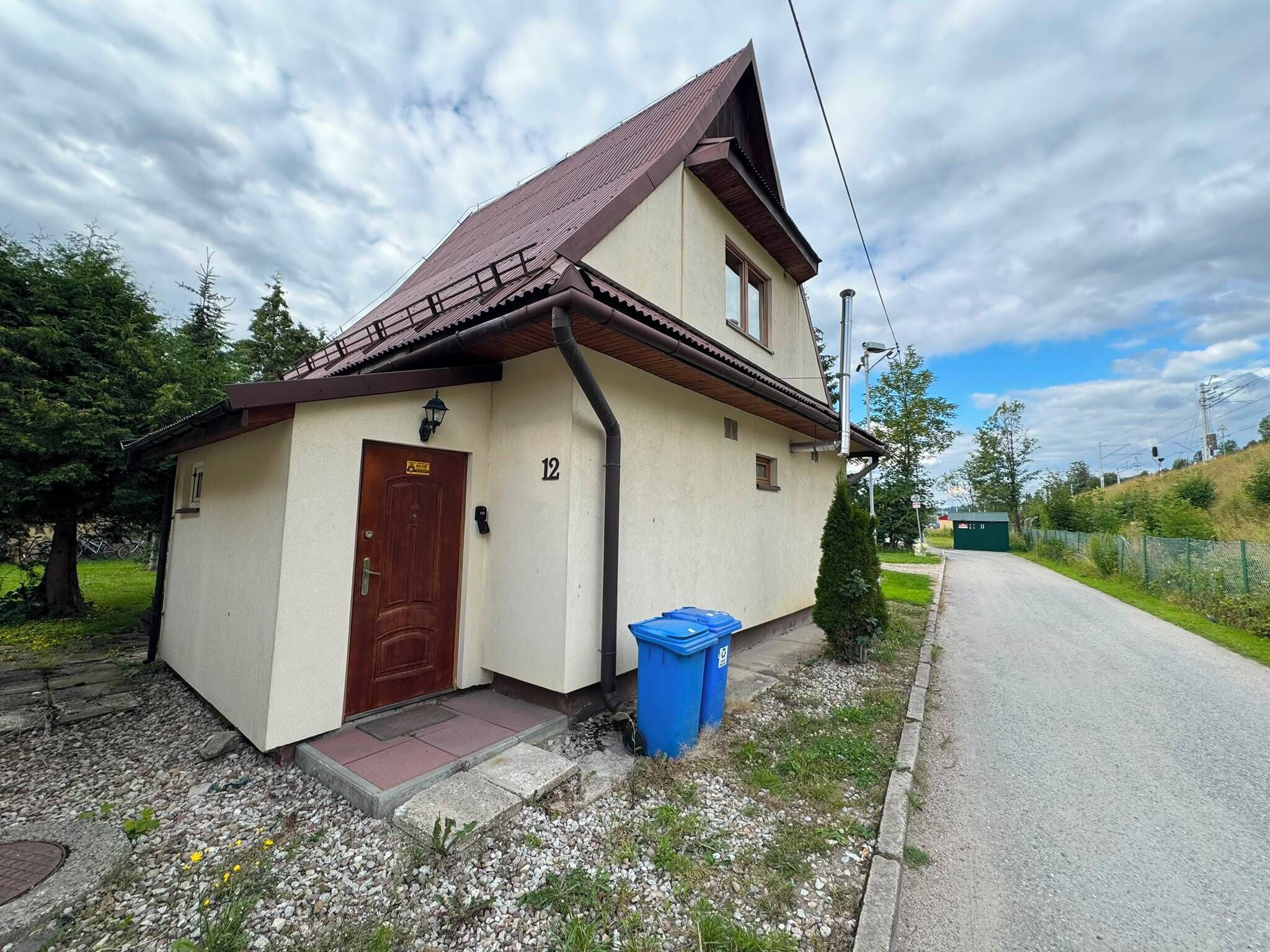 A single-story building with a brown door and a number '12' on the wall, a blue trash bin, and a gravel driveway.