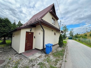A single-story building with a brown door and a number '12' on the wall, a blue trash bin, and a gravel driveway.