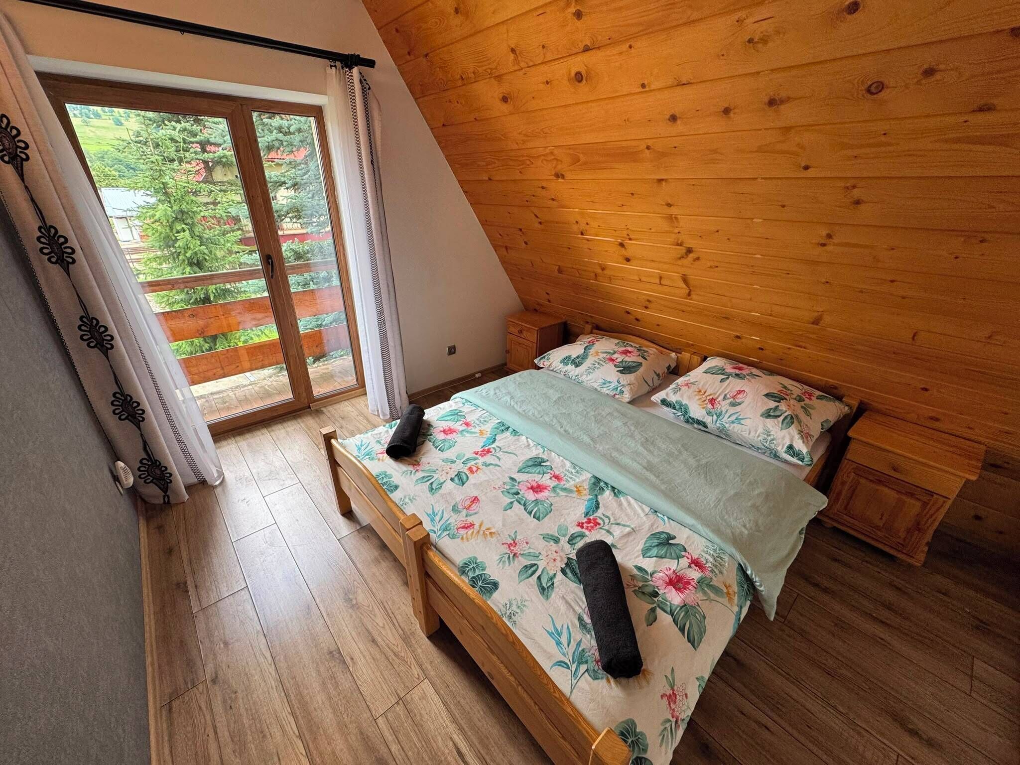 A bedroom with a wooden sloped ceiling, a bed with floral bedding, a wooden floor, and a sliding glass door leading to an outdoor area.