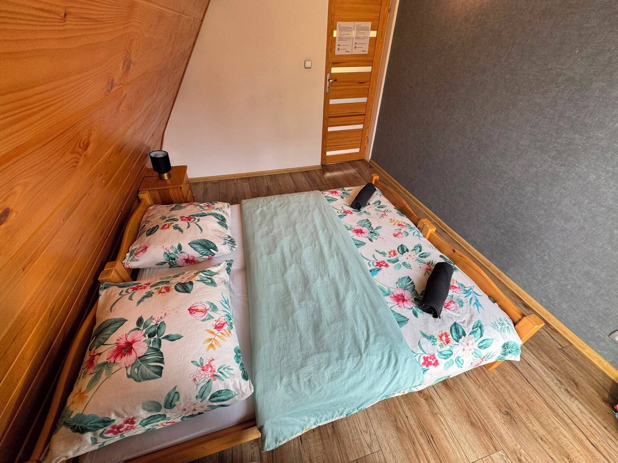 Un lit simple avec une literie à motifs floraux et une tête de lit en bois, dans une pièce dont le mur est gris et le sol en bois.