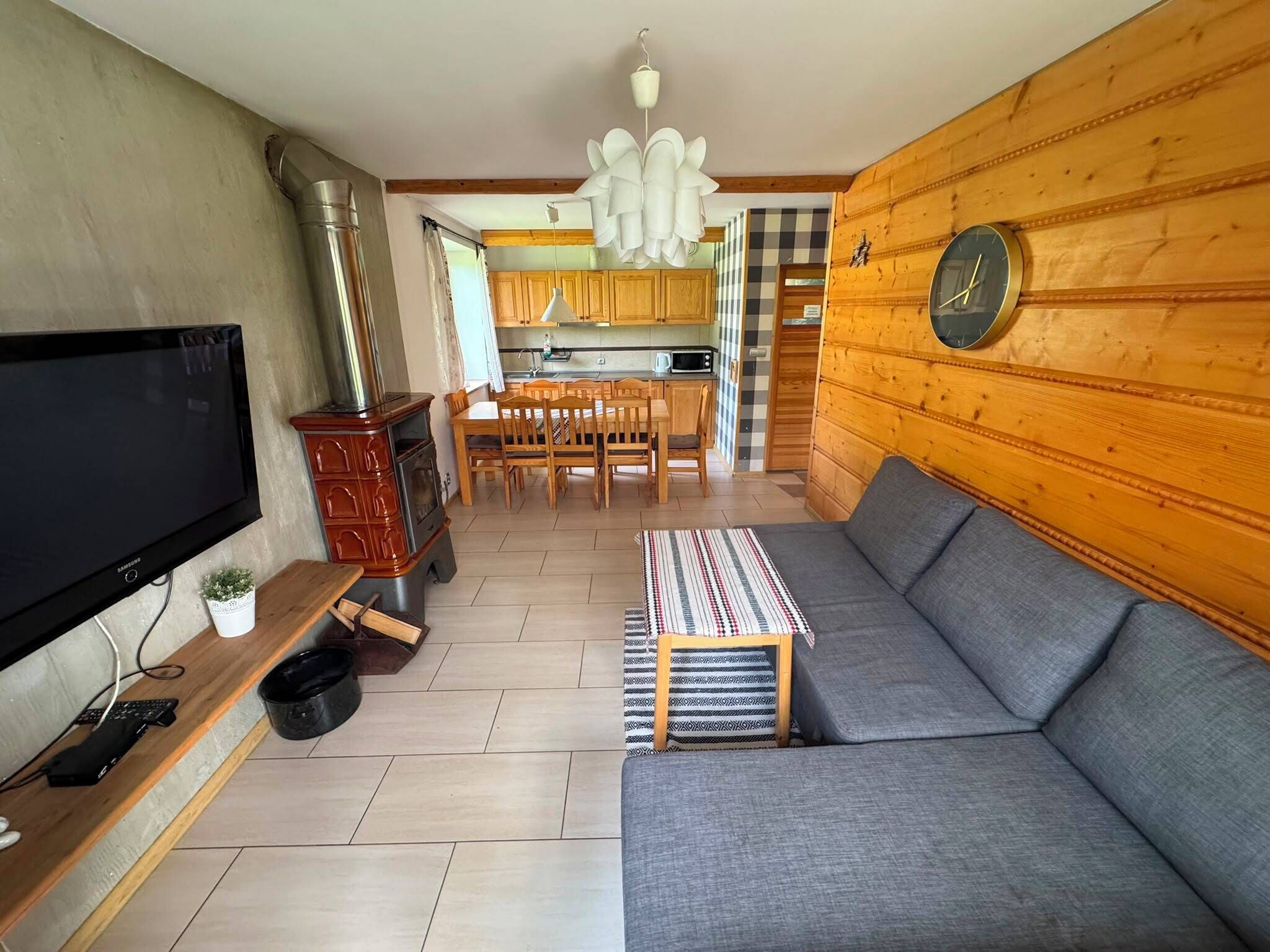 A living room with a grey sofa, wooden walls, a television, and a kitchen in the background.