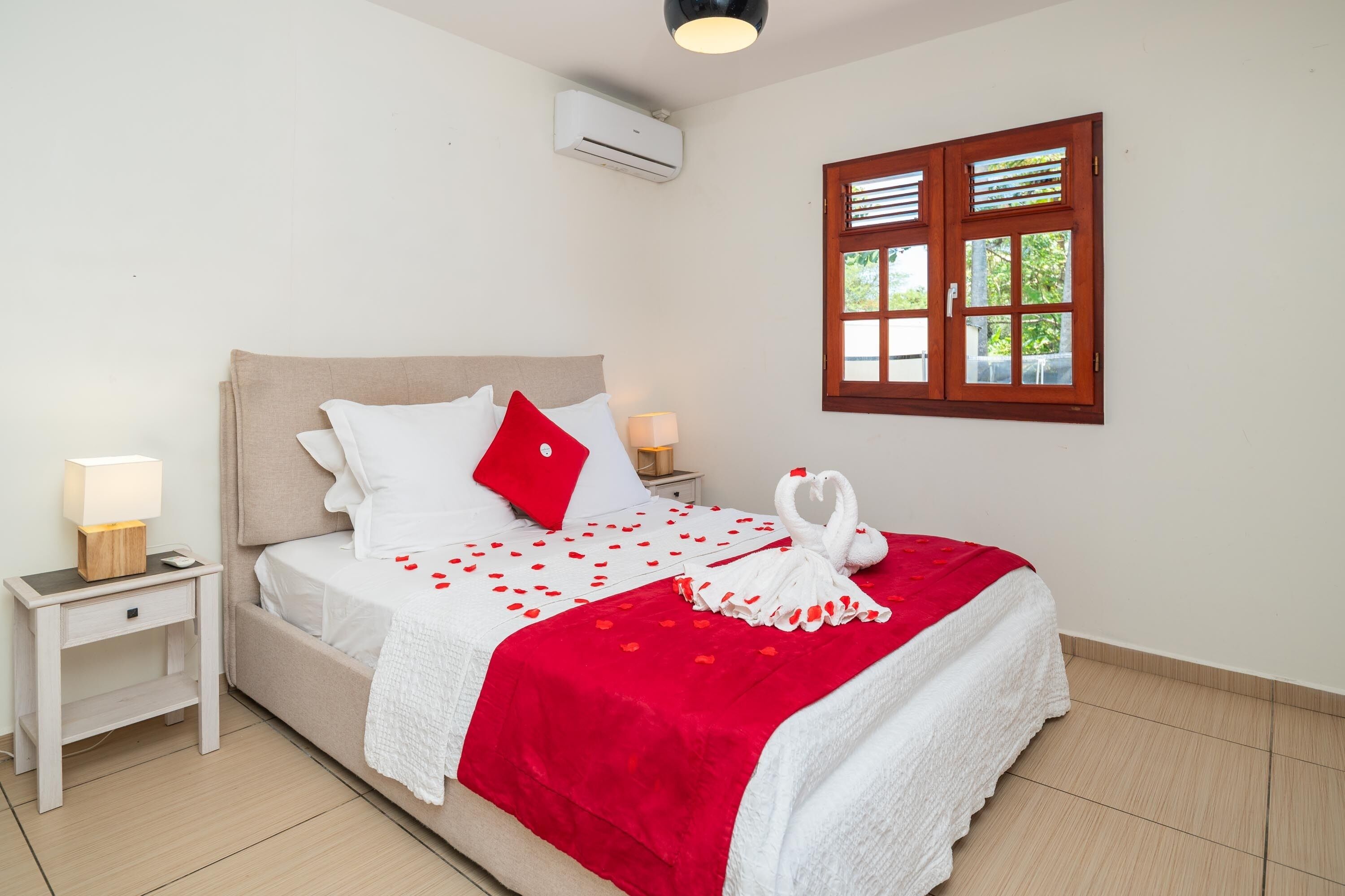 Una cama bien hecha con una colcha roja y blanca, dos lámparas de noche, una mesita de noche de madera y un aire acondicionado montado en la pared.