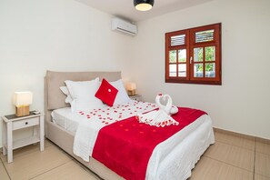 A neatly made bed with a red and white bedspread, two bedside lamps, a wooden nightstand, and a wall-mounted air conditioner. - L'Echappée Tropicale - Maison avec piscine privée (Le Moule)
