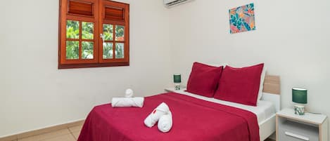 Habitación de hotel con cama, ventana con contraventanas, equipo de aire acondicionado, mesita de noche con lámpara y una obra de arte en la pared.