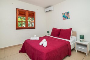 Quarto de hotel com cama, janela com persianas, aparelho de ar condicionado, mesa de cabeceira com abajur e uma obra de arte na parede.