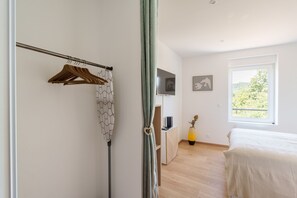Een slaapkamer met een bed, een televisie, een bureau met een computer en een raam met uitzicht op bomen.