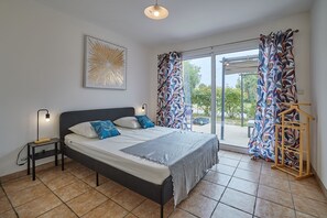 Un dormitorio con cama, una mesita de noche, lámpara, luz de techo, ventana con cortinas y vistas al exterior.