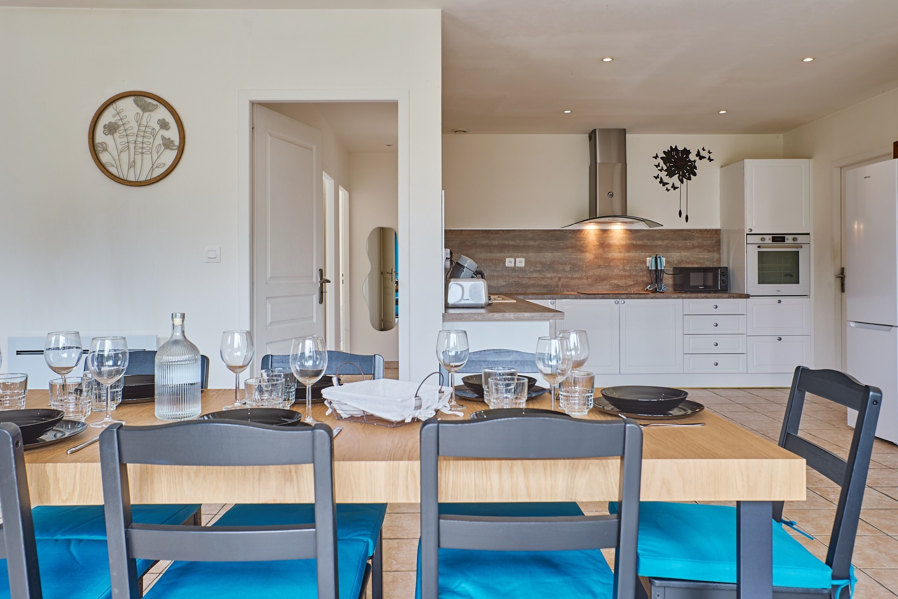 Un comedor con una mesa de madera dispuesta para cuatro, sillas y una cocina al fondo.