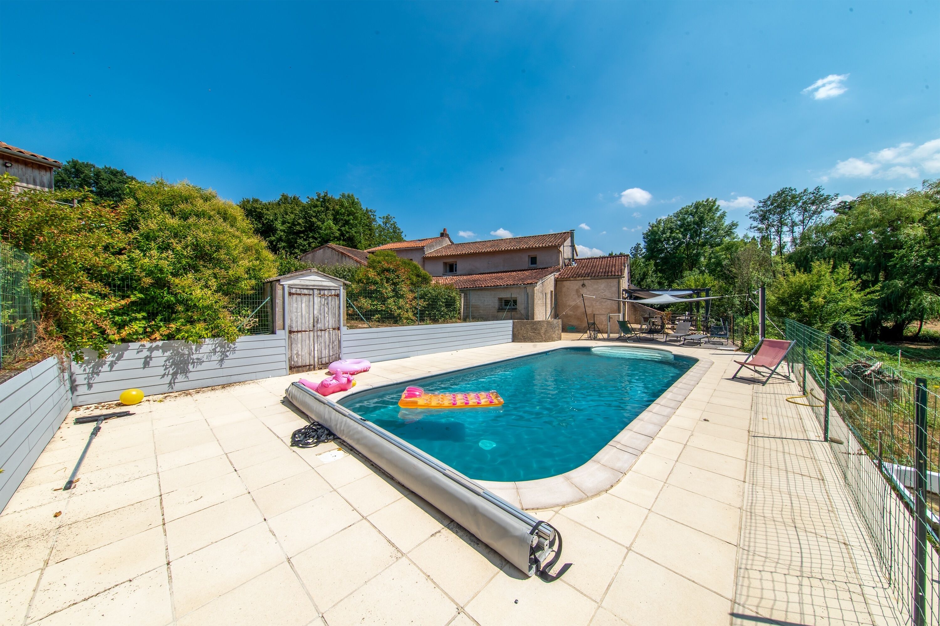Ein rechteckiger Pool mit gefliestem Rand, umgeben von einem Zaun und Bäumen.