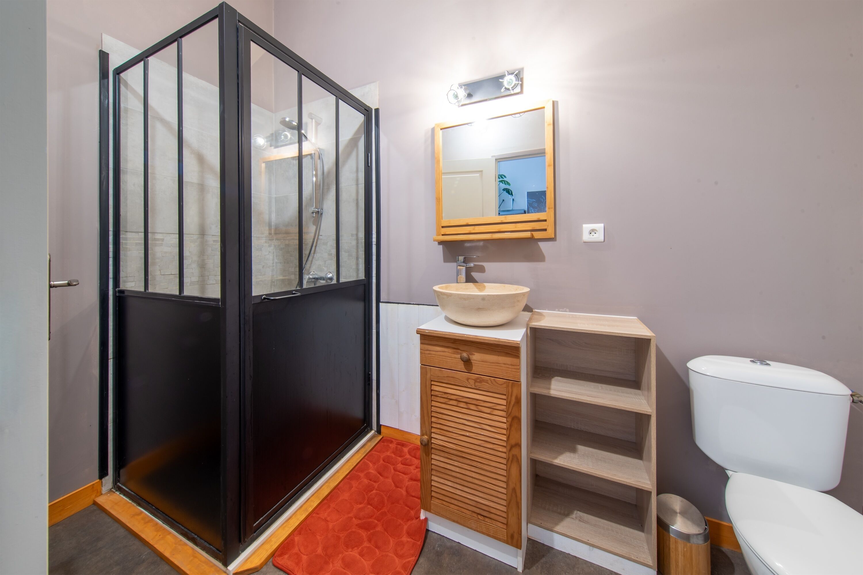 Un baño con cabina de ducha de vidrio, tocador de madera con lavabo, espejo y inodoro.