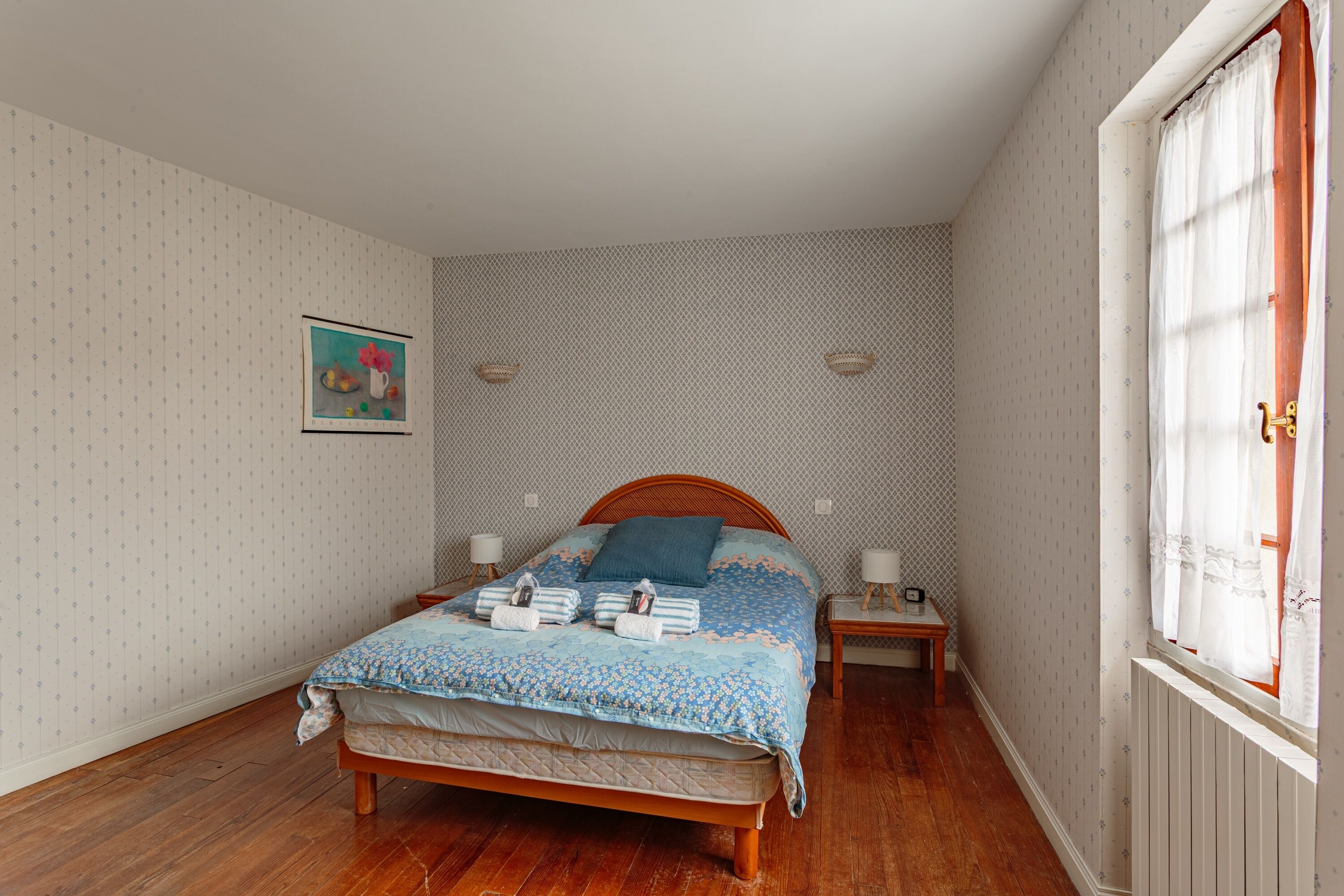 A bedroom with a wooden bed, a nightstand, a wall-mounted lamp, and a window with curtains.