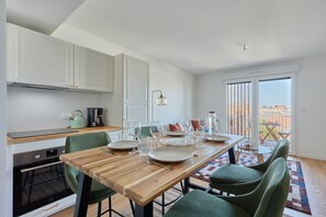 A modern kitchen with white cabinets, a wooden dining table set for four, and a view of the outdoors through a sliding glass door. - La Marina Dives - À 1 km de la plage (Dives-sur-Mer)