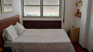 Un llit individual amb capçalera, una finestra amb persianes, un radiador i una estant amb llibres.