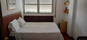 A single bed with a headboard, a window with blinds, a radiator, and a shelf with books.