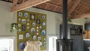 A cozy living room with a wooden ceiling, a fireplace, a wall decorated with blue and white plates, a chair with a fluffy pillow, and a small table with candles.