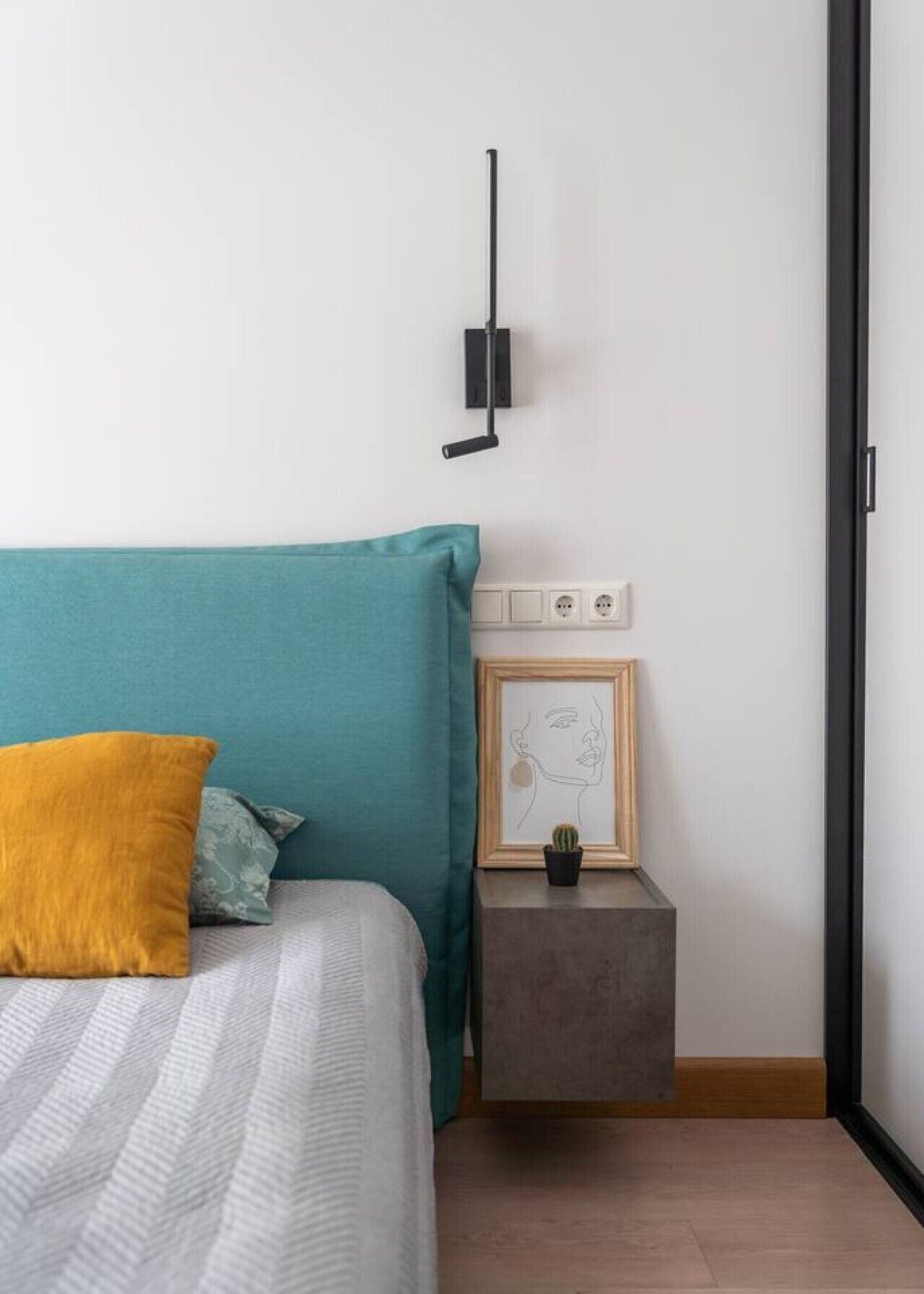Ein Schlafzimmer mit einem Bett, einem Nachttisch mit gerahmtem Bild, einer kleinen Topfpflanze und einer Wandleuchte.