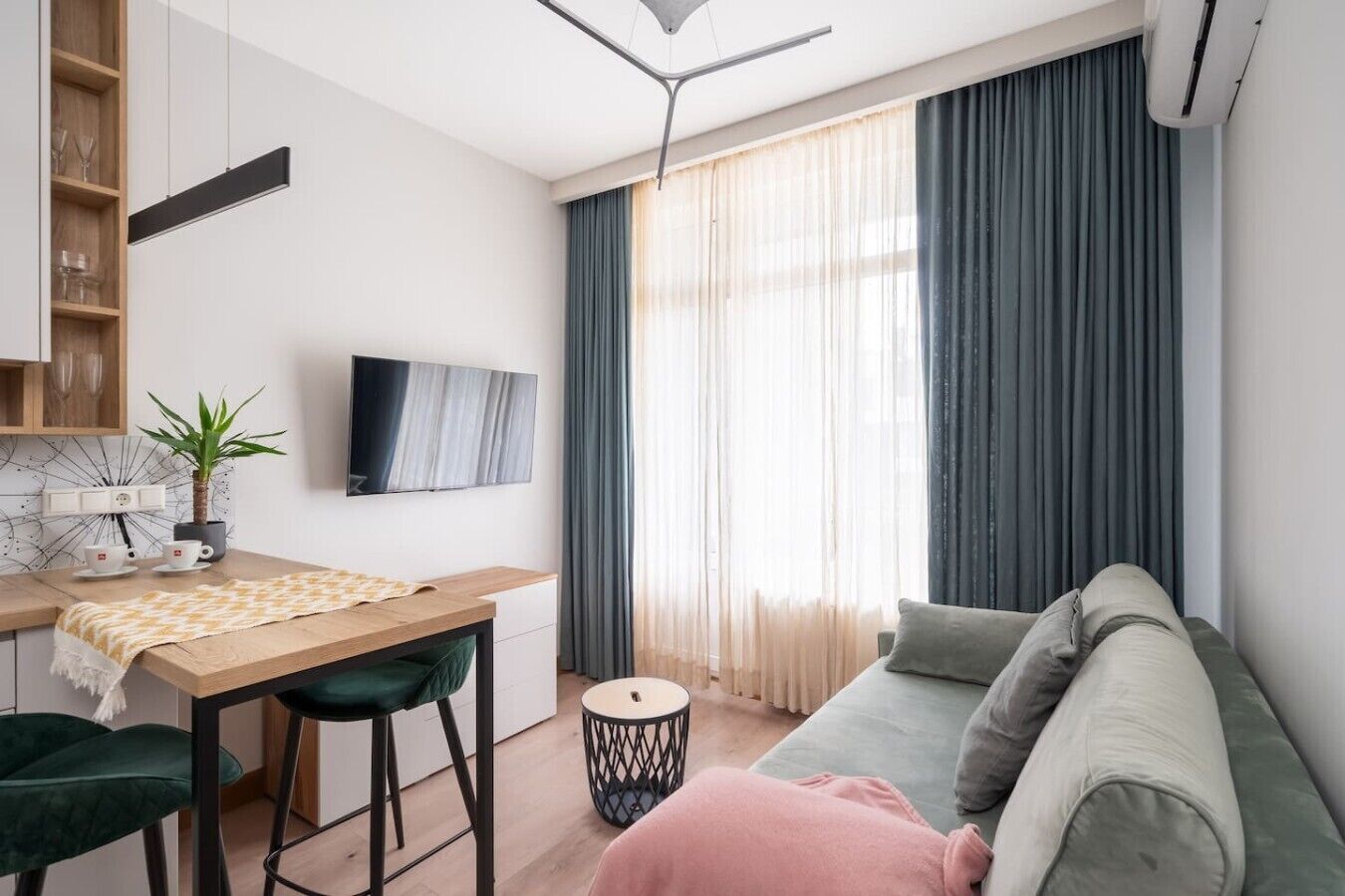 Uma sala de estar moderna com sofá, uma mesa de jantar com cadeiras, televisão na parede e uma janela com cortinas.