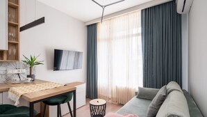 Uma sala de estar moderna com sofá, uma mesa de jantar com cadeiras, televisão na parede e uma janela com cortinas.