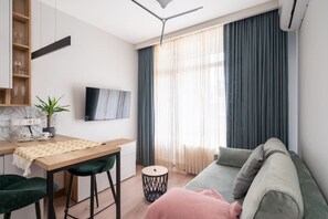 Ein modernes Wohnzimmer mit einer Couch, einem Esstisch mit Stühlen, einem an der Wand befestigten Fernseher und einem Fenster mit Vorhängen.