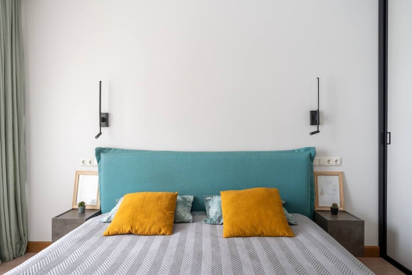 Ein Schlafzimmer mit einem türkisfarbenen Kopfteil, zwei Nachttischen mit Lampen sowie einem Bett mit einem gemusterten Bettbezug und zwei Kissen.