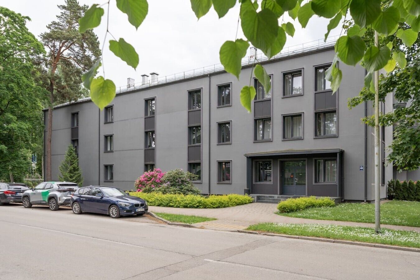 A modern building with multiple windows, a parking area with cars, and a well-maintained lawn with some flowering plants.