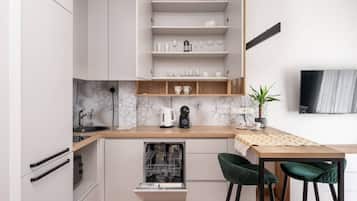 A compact kitchen with white cabinetry, a built-in dishwasher, a coffee maker, and a dining area with a wooden table and two green chairs.