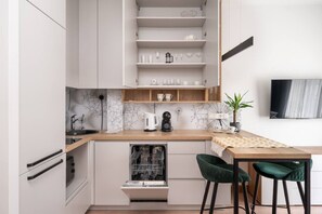 Une cuisine compacte avec des meubles blancs, un lave-vaisselle encastré, une machine à café et un coin repas comprenant une table en bois et deux chaises vertes.