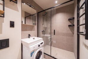 A modern bathroom with a built-in washing machine, a glass shower enclosure, and a wall-mounted sink. - Elegant 2-Bedroom 50m² Apartment | Old Town Riga (Riga)