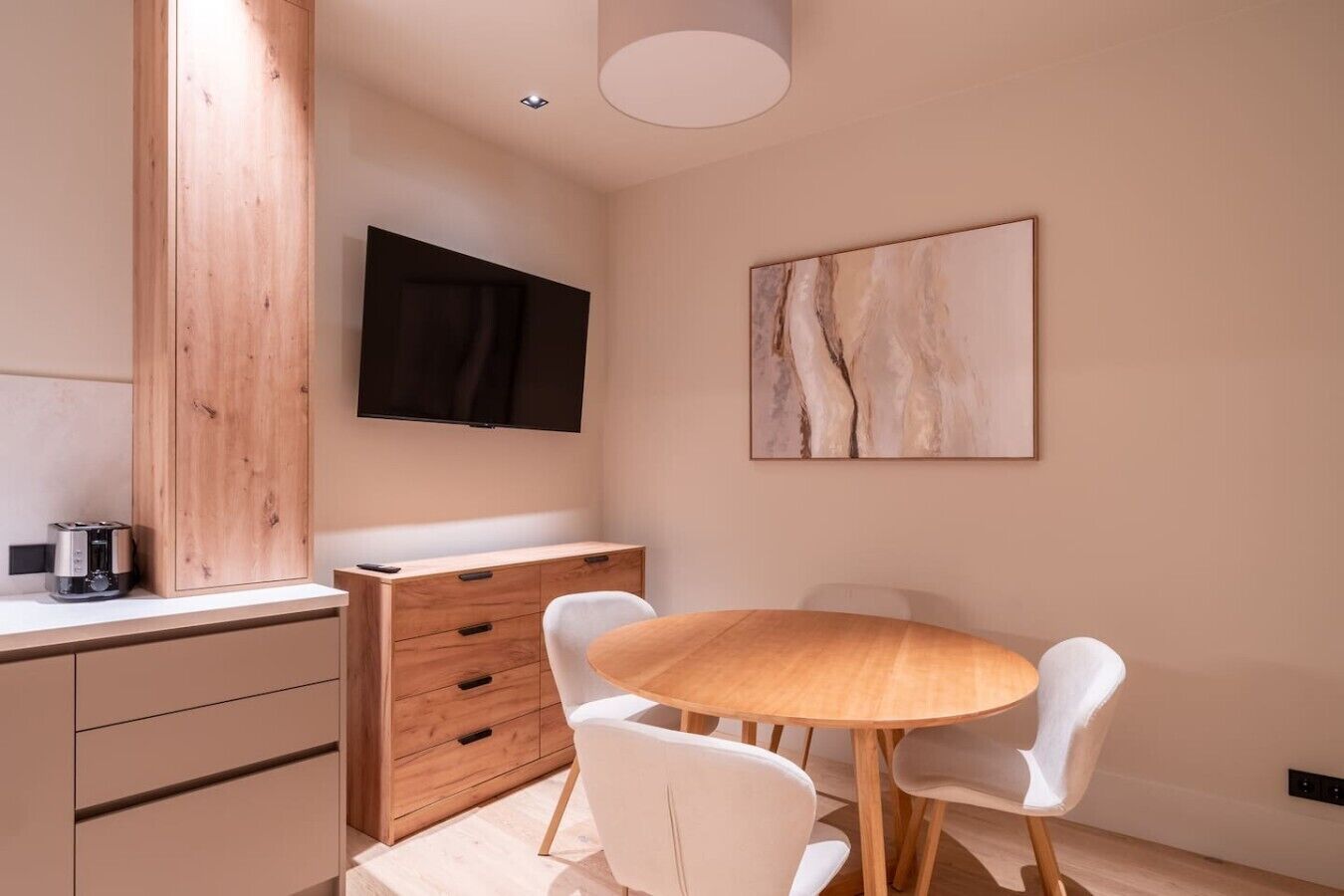 A modern kitchen with a wooden cabinet, a flat-screen TV, and a dining area with a round table and white chairs.