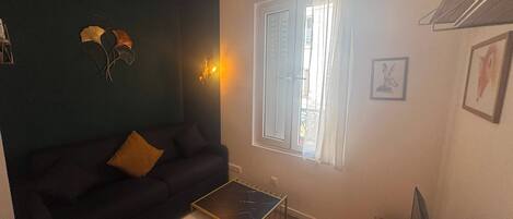 Un salon avec un canapé de couleur foncée, une petite table basse, une fenêtre avec des rideaux et un luminaire fixé au mur.