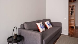 A grey sofa with two decorative pillows, a small black side table with a mesh object, and a wooden shelving unit in the background. - Spacious Family Haven in Riga’s Historic Heart (Rīga)