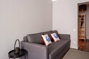 A grey sofa with two decorative pillows, a small black side table with a mesh object, and a wooden shelving unit in the background. - Spacious Family Haven in Riga’s Historic Heart (Rīga)