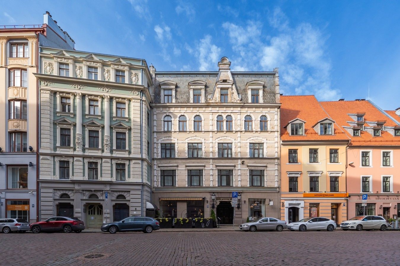 Une rangée de bâtiments historiques à l’architecture classique, une rue pavée et des voitures garées.